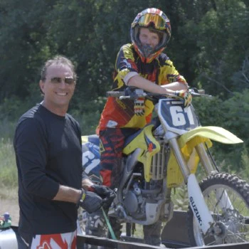 A person in a black shirt stands next to a motorcycle on a trailer, while another in motocross gear and helmet sits on it. Together, they represent the beginning of our story, set against the backdrop of an open outdoor adventure. – OmegaStrap – World’s Greatest Ratchet Strap A person in a black shirt stands next to a motorcycle on a trailer, while another in motocross gear and helmet sits on it. Together, they represent the beginning of our story, set against the backdrop of an open outdoor adventure. - OmegaStrap - World's Greatest Ratchet Strap