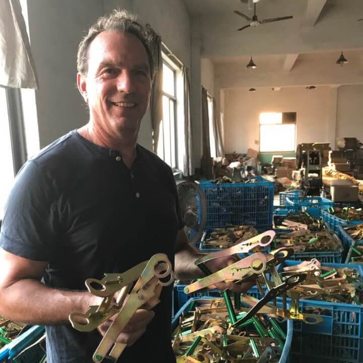 In a room filled with blue crates brimming with similar tools, a person holds metal instruments, echoing the beginnings of our story. – OmegaStrap – World’s Greatest Ratchet Strap In a room filled with blue crates brimming with ratchet strap parts, a person holds pieces, echoing the beginnings of our story. - OmegaStrap - World's Greatest Ratchet Strap