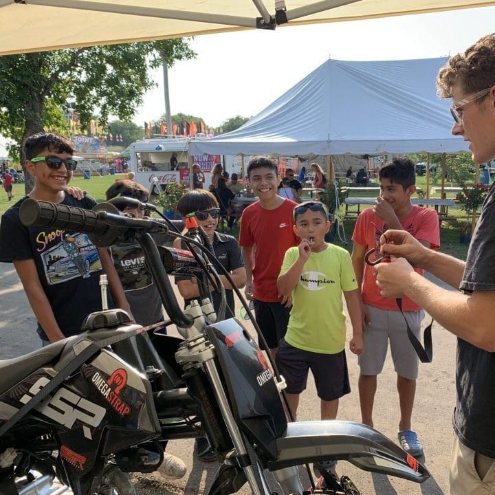 A group of boys gathers around a person showcasing a motorcycle with an OmegaStrap under a tent. The lively county fair buzzes in the background, complete with food stands and tables. – OmegaStrap – World’s Greatest Ratchet Strap A group of boys gathers around a person showcasing a motorcycle with an OmegaStrap under a tent. The lively county fair buzzes in the background, complete with food stands and tables. - OmegaStrap - World's Greatest Ratchet Strap
