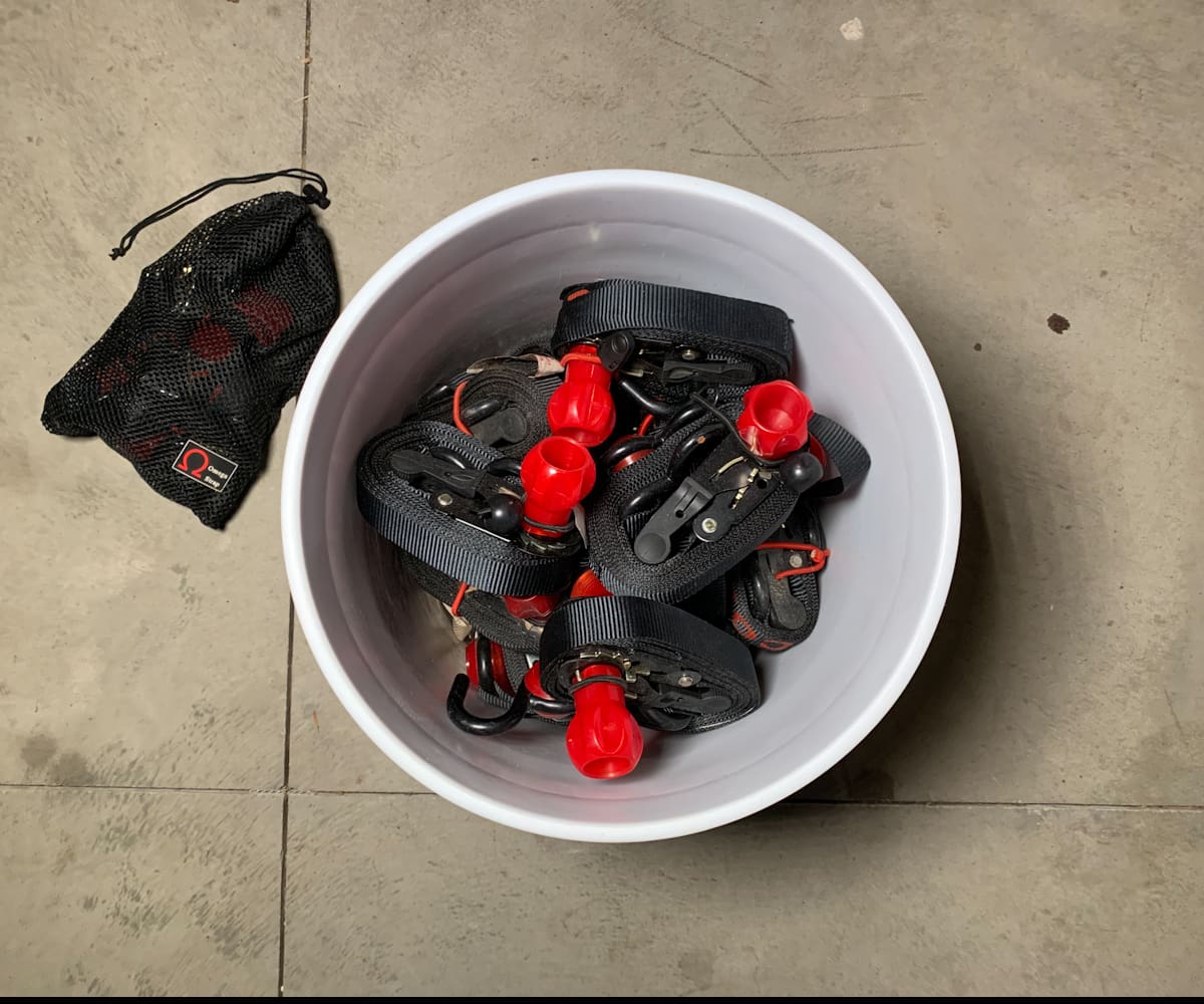 A white bucket containing several coiled OmegaStrap® ratchet straps with red handles is placed on a concrete floor next to a black mesh bag. - OmegaStrap - World's Greatest Ratchet Strap