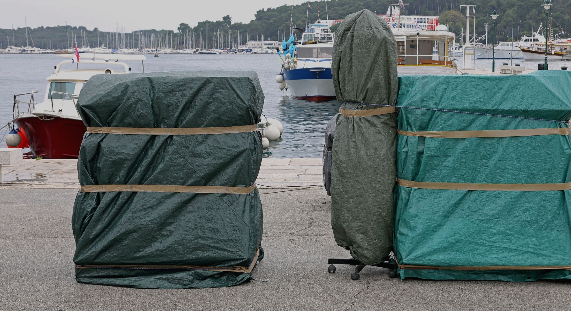 Two large objects covered with green tarps are secured on a dock using top ratchet strap accessories. Boats and a marina provide a scenic backdrop, perfectly complementing the efficient ratchet strap pairing that ensures everything stays in place. - OmegaStrap - World's Greatest Ratchet Strap